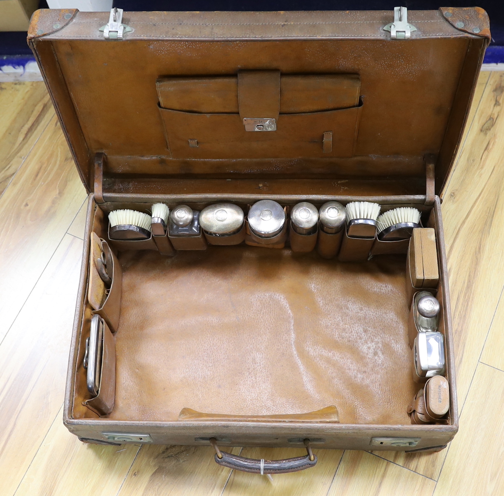 An Edwardian leather travelling toilet case, containing eleven silver mounted toilet jars etc. including a mounted glass hip flask and easel mirror, Drew & Sons, London, 1905, case width 62.5cm.